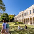 people walk and sit on grass by stone buildings with an arched walkway